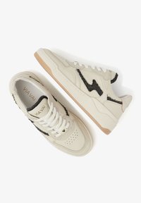 Beige sneakers with black patterned accents, featuring a textured finish, white laces, and a rubber sole with a light tan base.