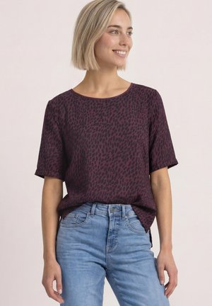 Ung kvinde med kort blondt hår, der smiler, iført en mørkelilla mønstret bluse og lyseblå jeans mod en ensfarvet baggrund.