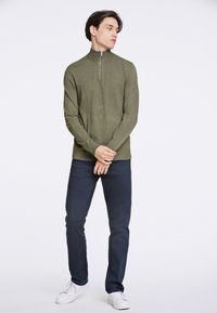 Olivgrüner Pullover mit Reißverschluss, taillierte Passform, gerippte Bündchen und Saum. Dazu dunkelblaue Slim-Fit-Hosen und weiße Sneakers. Schlichter Hintergrund.