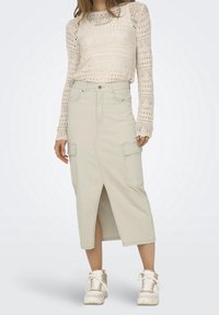 Jupe cargo beige clair avec une fente à l'avant, deux poches latérales, des passants pour ceinture et une fermeture à boutons. Portée avec un pull en tricot crème et des baskets blanches.