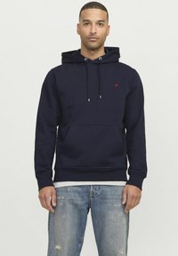 Hoodie en coton bleu marine avec une poche avant, capuche à cordon et petit logo rouge sur la poitrine. Porté sur une chemise gris clair et un jean déchiré.
