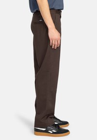 Brown cotton trousers with a straight leg design, featuring a back pocket and worn with black sneakers and a blue shirt.