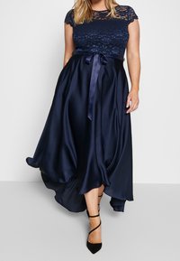 Sini sininen satiinimekko, jossa on pitsinen yläosa, cap-sleeve-hihat ja tiukka vyötärö, joka on sidottu satiinivyvyn. Epäsymmetrinen hameen muotoilu. Mustat korkokengät täydentävät asua.