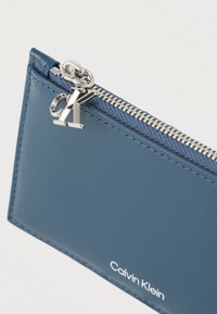 Πορτοφόλι σε ναυτικό μπλε με φερμουάρ, κατασκευασμένο από λείο δέρμα, με ασημένιο μενταγιόν λογότυπο και τυπωμένο όνομα μάρκας στο μπροστινό μέρος.