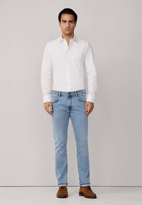 Biała koszula na guziki, jasnoniebieskie dżinsy, brązowe buty. Koszula ma gładką fakturę, a dżinsy posiadają standardowe kieszenie i delikatne przetarcia.