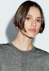 Cardigan en maille gris avec un col rond, un tissu texturé et une fermeture à deux boutons. Cheveux courts et grandes boucles d'oreilles en argent visibles.