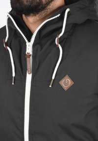 Sort jakke med lynlås foran, hvide snore og et logo-patch i brun læder. Har en glat tekstur og en justerbar hætte.