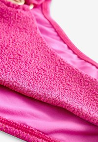 Růžový bikinový top s texturovanou tkaninou se třpytem, kontrastní hladkou vnitřní podšívkou a zaobleným výstřihem s detaily na zapínání.