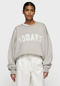 Felpa grigia oversize con il testo bianco "10DAYS", scollo rotondo, polsini a costine e un morbido tessuto testurizzato. Indossata sopra pantaloni bianchi.