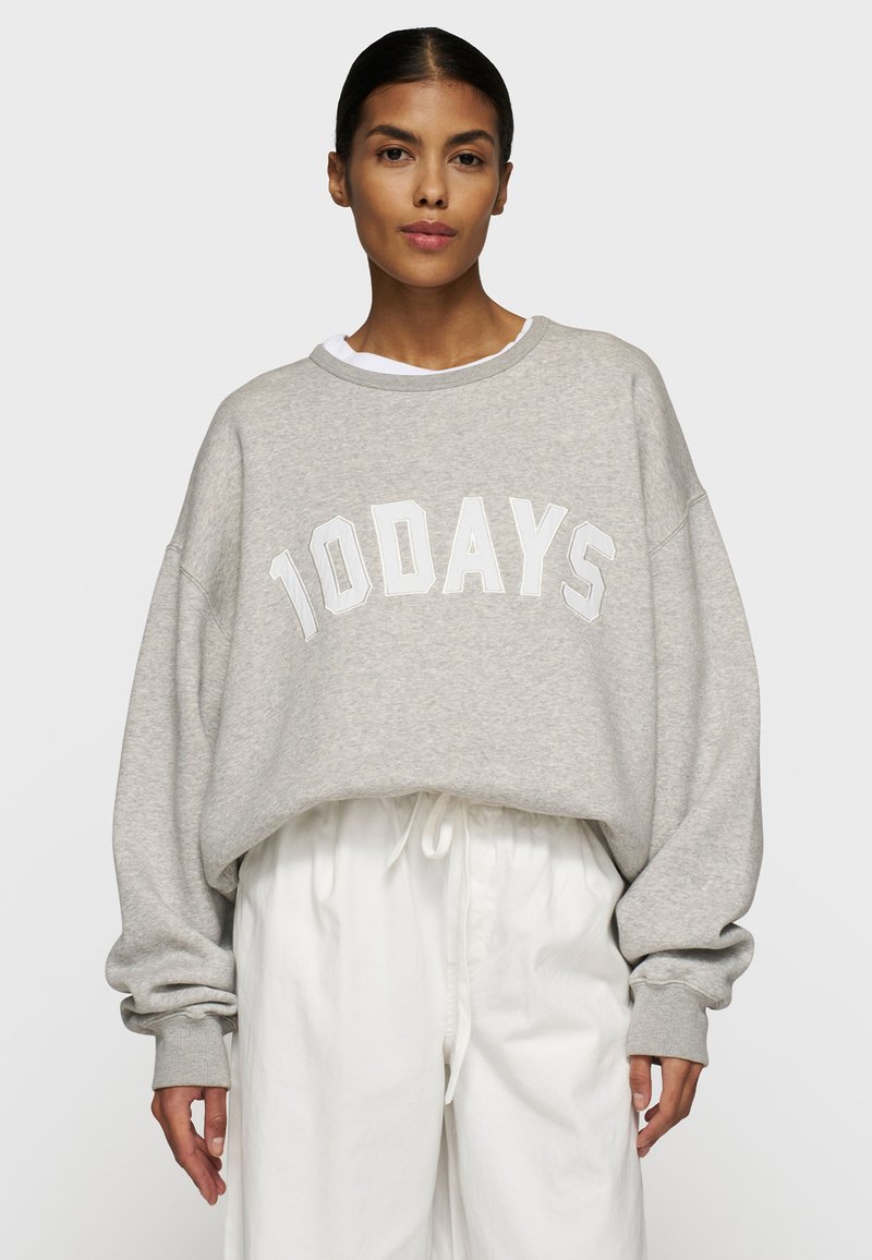Felpa grigia oversize con il testo bianco "10DAYS", scollo rotondo, polsini a costine e un morbido tessuto testurizzato. Indossata sopra pantaloni bianchi.
