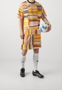 Oranssi ja keltainen kuvioitu urheilu-paita ja shortsit, mustat korostusviivat, yhdistettynä mustiin jalkapallokenkiin ja jalkapalloon.