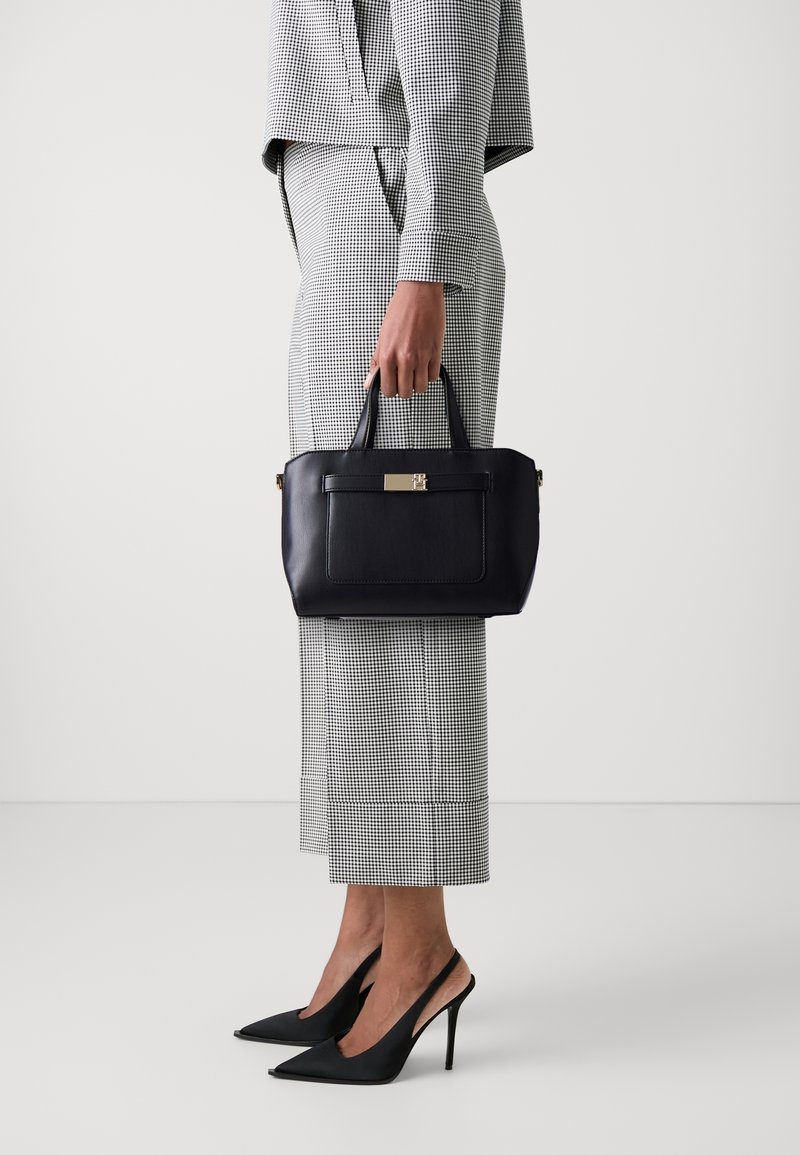Personne en costume à carreaux noir et blanc tenant un sac à main en cuir noir, portant des chaussures slingback noires à talons aiguilles pointus.