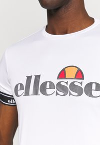 Bijela majica kratkih rukava s velikim crnim "ellesse" logotipom sa šarenom grafikom iznad. Crne prugaste akcenti na rukavima.