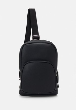 Kompakt sort crossbody taske med justerbar rem, front lynlåslomme og tekstureret materiale på en ensfarvet lys baggrund.