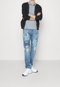 Ljusgrå pikétröja under en svart kofta, kombinerad med slitna blå jeans med lapptäckesdetaljer och vita sportskor.