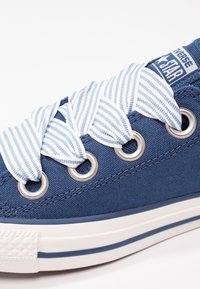 Baskets en toile bleue avec semelle en caoutchouc blanche. Elles présentent des lacets rayés bleu et blanc, des œillets en métal et une étiquette sur la languette.