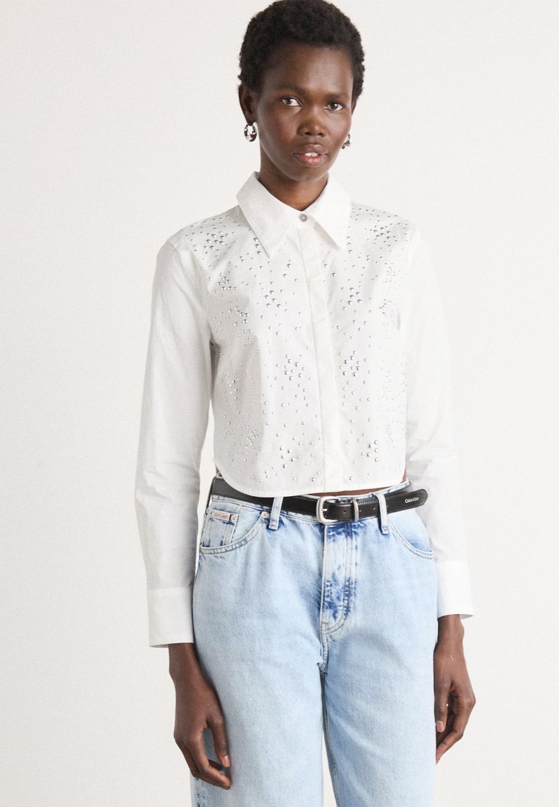 Camicetta bianca corta con dettagli ricamati e bottoni, abbinata a jeans in denim azzurro chiaro e una cintura nera. Tessuto liscio e colletto strutturato.