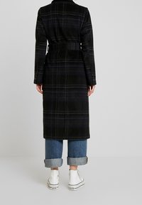 Persona in piedi contro uno sfondo chiaro, vestita con un lungo cappotto di tessuto a quadri scuri con cintura, jeans blu risvoltati e sneakers alte bianche Converse.