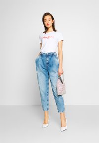 T-shirt bianca con testo ricamato rosso, jeans a vita alta strappati di un blu chiaro e una piccola borsa scintillante. Le scarpe sono bianche.