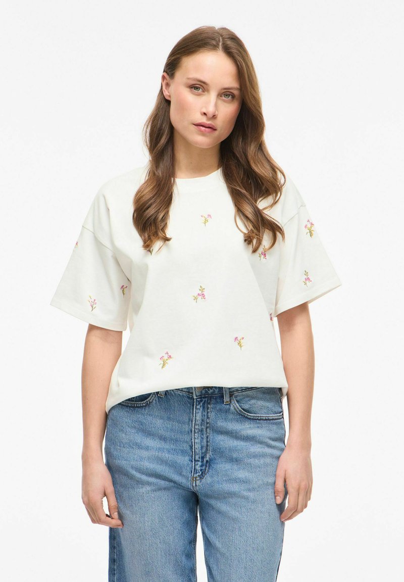 Femme aux longs cheveux bruns portant un t-shirt blanc oversize avec de petites fleurs roses imprimées et un jean bleu, debout devant un fond blanc uni.