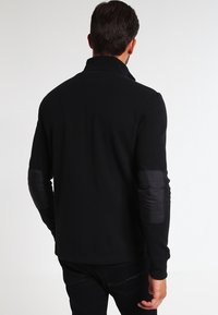 Černý svetr s texturovaným úpletem a stojáčkovým límcem. Má černé látkové náplasti na loktech pro zvýraznění detailu. Moderní střih a design.