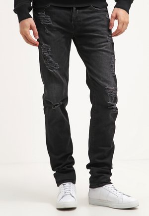 Mężczyzna w czarnych podniszczonych dżinsach i białych tenisówkach, stojący na jednolitym jasnym tle.