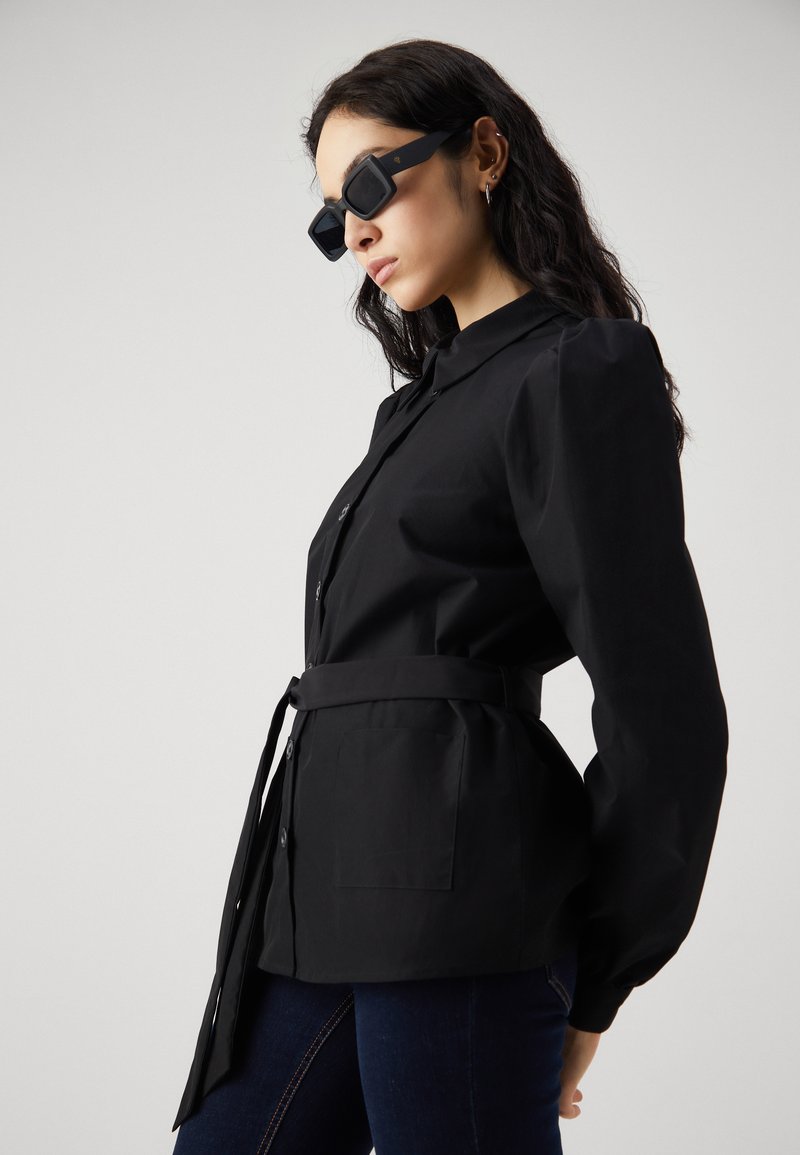 Schwarze Hemdbluse mit Puffärmeln, tailliertem Schnitt und zwei vorderen Taschen. Tragen mit schwarzen quadratischen Sonnenbrillen und dunklen Jeans.