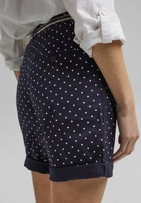 Personne portant un short bleu marine à pois blancs avec ourlets retroussés, associé à une chemise blanche à manches longues retroussées.