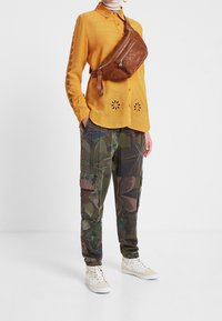 Camicia gialla con bottoni e intagli floreali, abbinata a pantaloni cargo mimetici verdi con tasche. Marsupio marrone e sneakers bianche con motivi floreali.
