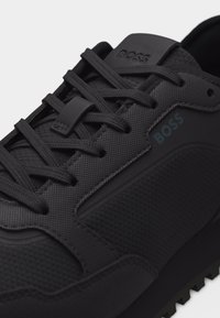 Μαύρο κλωστοϋφαντουργικό αθλητικό παπούτσι με μαύρα κορδόνια, με την επωνυμία "BOSS" στη γλώσσα και στην πλευρά, με ανθεκτική σόλα.