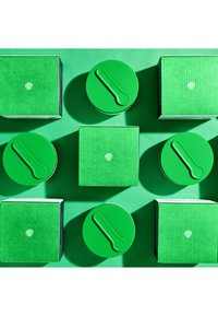 Grüne Verpackung mit strukturierter Oberfläche. Enthält runde Behälter mit flachen Deckeln und rechteckige Schachteln, die in einer symmetrischen Anordnung platziert sind.