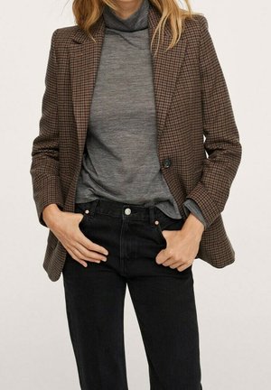 Donna che indossa un blazer marrone a quadri sopra a un maglione dolcevita grigio, infilato in jeans neri a vita alta con le mani nelle tasche.