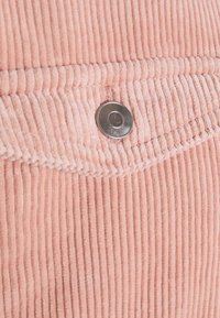 Różowy materiał sztruksowy z pionowymi żebrowaniami, z metalowym zatrzaskiem u góry i gładką linią szwu.