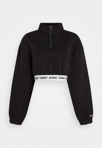 Sudadera corta negra con cuello alto, cierre de media cremallera en el frente y mangas anchas. Presenta una banda elástica blanca con marca en la cintura.