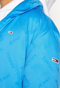 Light blue puffer jacket featuring a hood, textured fabric, and all-over printed "TOMMY JEANS" pattern; includes a small logo patch on sleeve.