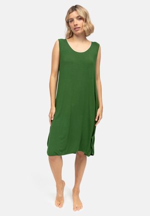 Frau steht barfuß und trägt ein ärmelloses, locker sitzendes grünes knielanges Kleid vor einem schlichten hellen Hintergrund.