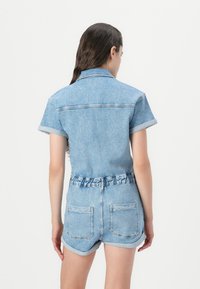 Salopette en denim bleu clair avec manches courtes, taille ceinturée et poignets roulés. Comprend deux poches arrière et un col.