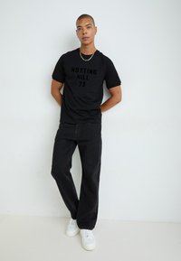 Svart bomull T-skjorte med "NOTTING HILL 73" i hevet svart tekst, sammen med svarte jeans og hvite sneakers, satt mot en lys bakgrunn.