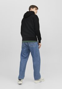 Zwarte hoodie, groene shirt eronder, lichtblauwe baggy jeans, sneakers met rubberen zool, en een neutrale achtergrond.