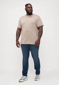 Bež pamučna majica s printom logotipa, uparena s plavim trapericama. Model nosi bijele i sive tenisice. Jednostavan, ležeran outfit.