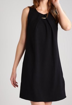Kobieta w czarnej, bezrękawnikowej sukience do kolan z małym złotym paskiem blisko dekoltu na jednolitym tle.