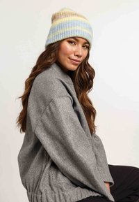 Knit beanie in pastel yellow, blue, and tan stripes; soft texture. Worn with a gray, oversized sweater featuring a cinched waistband.