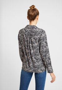 Black and cream patterned shirt with long sleeves, collar, and back pleats. Paired with blue jeans, the shirt features a fluid texture.
