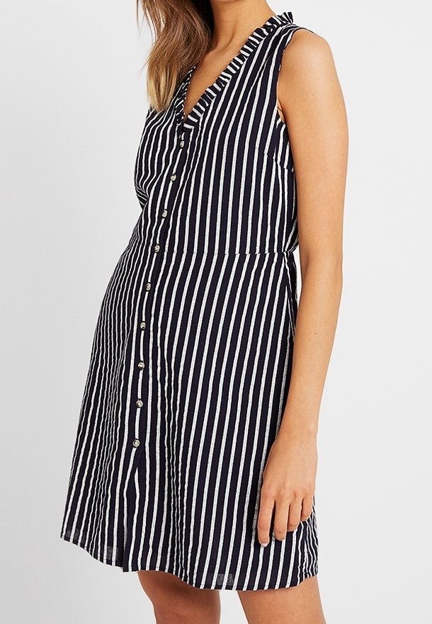 Robe sans manches rayée verticalement noire et blanche, avec des boutons sur le devant et une encolure à volants, portée par une personne aux longs cheveux.