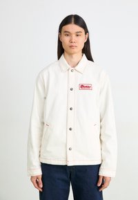 Jachetă din denim albă cu închidere cu nasturi, detalii cu cusături roșii și un petic pătrat roșu cu text alb. Model purtând blugi de culoare albastră închisă.