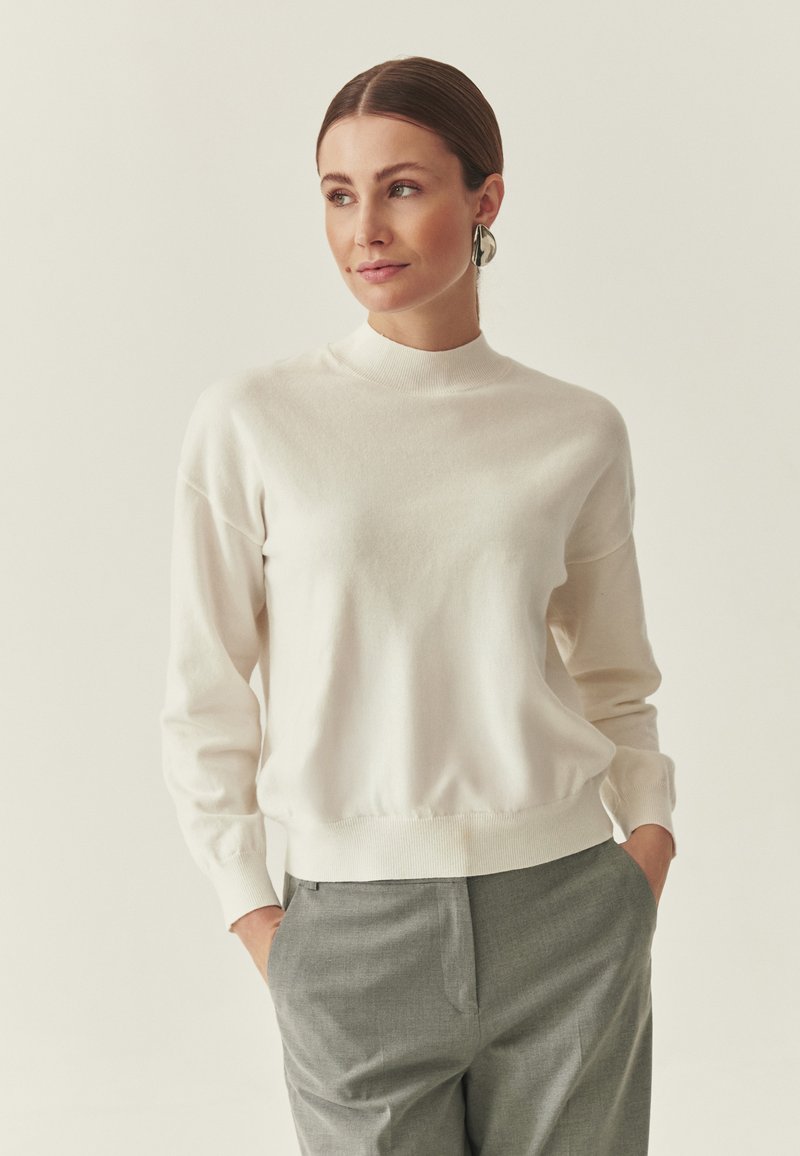 Pull en tricot crème avec col montant, manches longues et poignets côtelés, assorti à un pantalon gris taillé. Texture douce, design minimaliste.