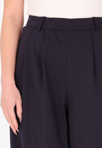 Pantaloni blu a vita alta con pieghe, realizzati in materiale morbido. Presentano passanti per cintura e una texture liscia, visibile sulla vita.