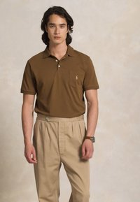 Moški s srednje dolgimi temnimi lasmi, oblečen v rjavo polo majico in bež hlače s pregibi, stoji pred nevtralno ozadjem.