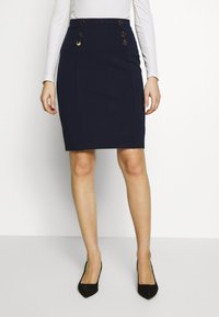 Námořnická tužková sukně vyrobená z pružné tkaniny, s čtyřmi zlatými knoflíkovými akcenty na přední straně. Hladká textura s přiléhavým střihem.