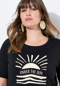 Camiseta negra de cuello redondo con un gráfico en crema de un sol naciente y líneas onduladas, combinada con grandes pendientes circulares de mimbre.